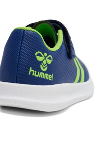Blå sneaker med en texturerad yta, gröna accenter och en vit sula. Har en kardborreband och Hummel-logotyp.