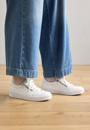 Persoon draagt lichtblauwe wijde jeans en witte leren sneakers met zijritsen, staat op een houten vloer.