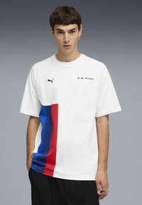 Νέος άνδρας που φοράει λευκό μπλουζάκι Puma με λογότυπα BMW Motorsport και κάθετες μπλε και κόκκινες ρίγες, στέκεται μπροστά σε γκρι φόντο.