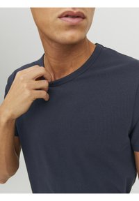 Muž v tmavě modrém tričku s kulatým výstřihem, který si pravou rukou mírně tahá za límec, na jednostranném světlém pozadí.