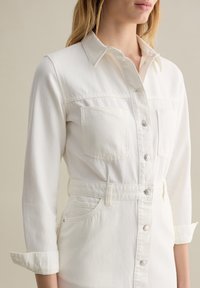 Abito in denim bianco con colletto, chiusura frontale a bottoni, due tasche sul petto e maniche arrotolate, caratterizzato da una vita aderente e una consistenza liscia.