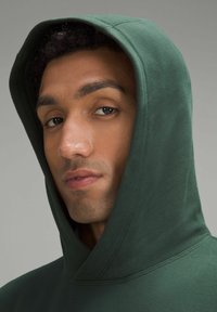 Groene hoodie met een grote capuchon, gemaakt van zachte stof. Close-up van het gezicht van de persoon, met een focus op de gladde huid en het donkere haar.