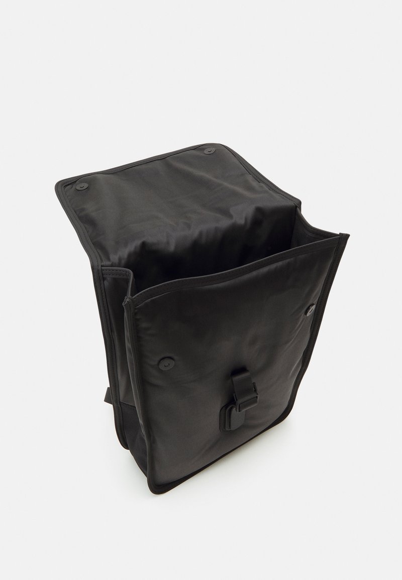 Zaino nero con una texture morbida, parte superiore con patta, chiusure a bottone e spallacci regolabili. L'interno è spazioso e non foderato.