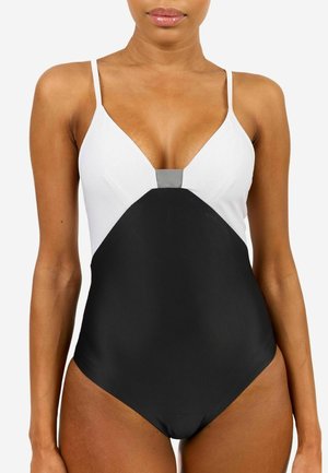 Woman wearing a black and white one-piece swimsuit with thin shoulder straps and a deep V neckline against a white background.