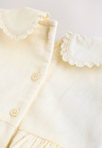 Dos prendas de bebé color crema con cuellos de encaje con borde festoneado y pequeños botones sobre tela suave.