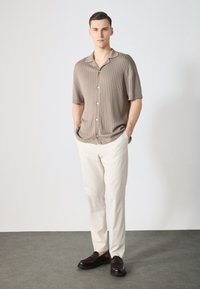 Beige pruhovaná košile s krátkým rukávem a knoflíky, spárovaná s bílými kalhotami a tmavě hnědými mokasíny, na modelu na světlém pozadí.