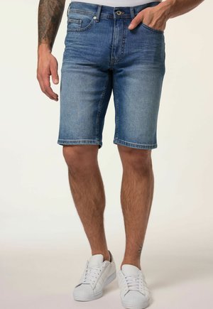 Homme portant un short en jean bleu et des baskets blanches, debout avec une main dans la poche devant un fond uni.