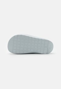 Suela de zapatilla de goma blanca con un patrón de cuadrícula texturizado, detalle sutil del logo y bordes suaves. Acabado suave con una ligera curvatura en la punta.