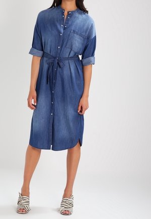 Kobieta w niebieskiej dżinsowej sukience koszulowej do połowy uda z podwiniętymi rękawami, wiązanym w talii paskiem, zapinanej z przodu na guziki, w paskowane sandały na obcasie z odkrytymi palcami.