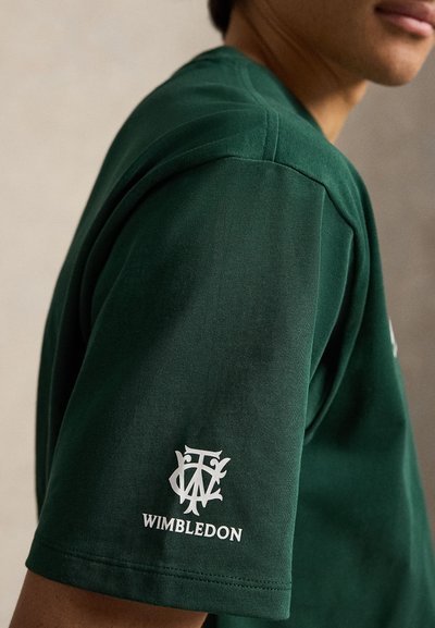 Άτομο που φοράει σκουρό πράσινο T-shirt με λευκό λογότυπο του Γουίμπλεντον στο μανίκι, εμφανιζόμενο από τον ώμο έως το μέσο του κορμού σε ουδέτερο φόντο.