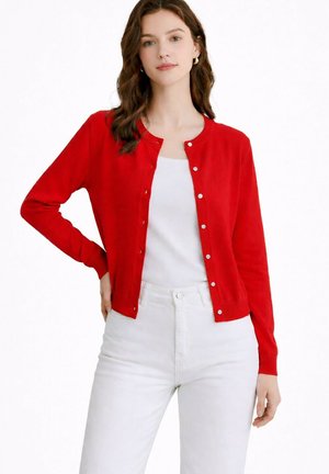 Femme portant un cardigan rouge boutonné sur un haut blanc et un pantalon blanc, debout avec une main sur la hanche devant un fond blanc.