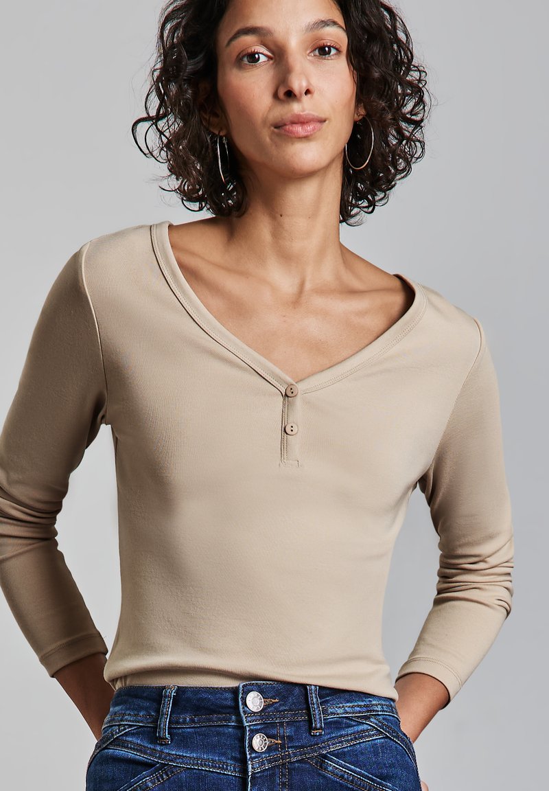 Haut beige à manches longues avec un décolleté en V et deux boutons décoratifs. Tissu doux avec une coupe ajustée. Associé à un jean bleu foncé.
