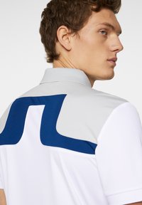 White polo shirt with a gray collar and a large blue geometric design on the back. Features short sleeves and a smooth fabric texture.