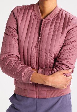 Person wearing a dusty pink quilted bomber jacket with ribbed cuffs and collar, arms crossed, paired with purple pants.