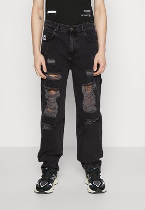 Persoon draagt zwarte distressed jeans met grote scheuren op de dijen en zwart-witte sneakers, staand tegen een witte achtergrond.