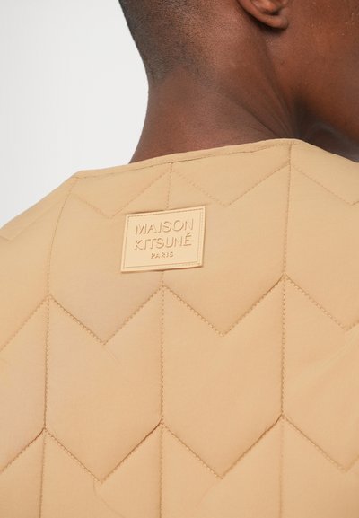 Tessuto tan trapuntato con un motivo a diamante, caratterizzato da un'etichetta in pelle rettangolare impressa con "MAISON KITSUNÉ PARIS" all'altezza del collo.