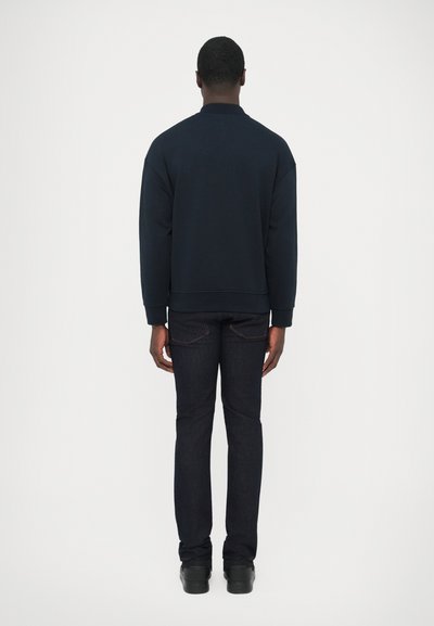 Tmavomodrá mikina s okrúhlym výstrihom a rebrovaným lemom, skombinovaná s tmavými denimovými džínsami. Jednofarebný materiál s hladkou textúrou bez viditeľných vzorov.