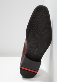 Semelle d'une chaussure habillée en cuir marron avec un motif de semelle hexagonal et un accent rouge sur le talon noir, positionnée verticalement contre une surface blanche.