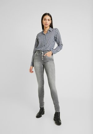 Camisa de botão xadrez azul-marinho e branca, combinada com jeans cinzentos de cintura alta e botins pretos, realçando uma silhueta elegante.