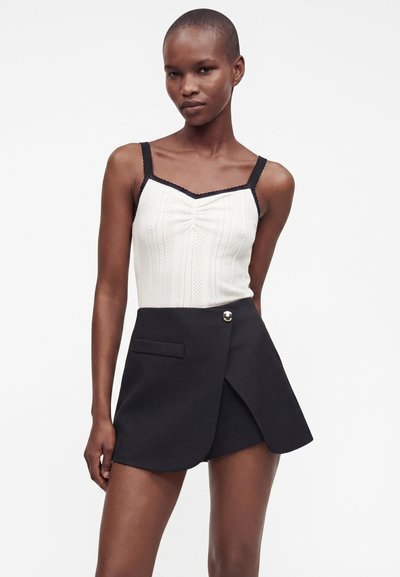 Femme portant un haut blanc sans manches avec des bretelles noires et une jupe portefeuille noire taille haute avec détail bouton.