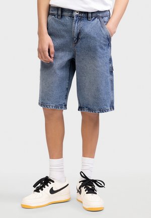 Νεαρός που φορά μπλε τζιν σορτς, λευκές κάλτσες και λευκά αθλητικά παπούτσια με μαύρα λογότυπα Swoosh, στέκεται με τα χέρια κοντά στις τσέπες.