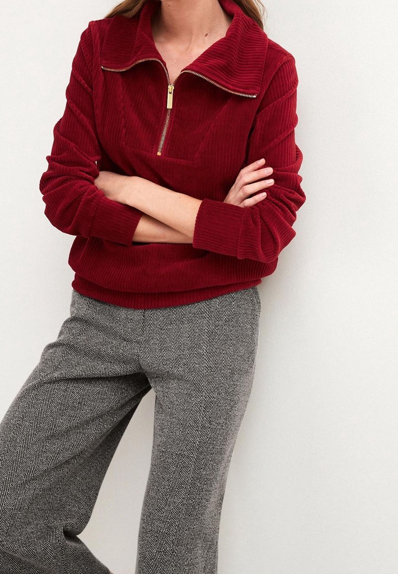 Pull en velours côtelé bordeaux avec un col haut et une fermeture éclair dorée. Associe à un pantalon en chevron gris. Texture douce, coupe décontractée.