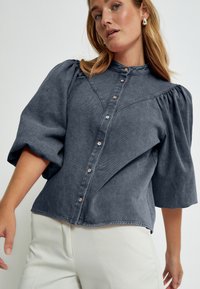 Denimbluse in Dunkelgrau, mit Puffärmeln, durchgehender Knopfleiste und spitzen Kragen. Subtile Textur mit dekorativen Nähten.