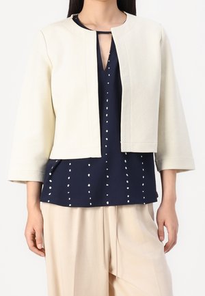Femme portant une veste courte crème ouverte sur le devant, par-dessus un chemisier bleu marine à pois blancs verticaux et un pantalon beige ample.
