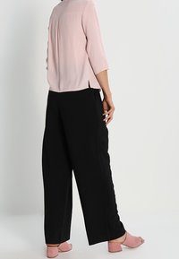 Blouse rose clair à manches trois-quarts avec pli au dos, associée à un pantalon noir à jambes larges et des chaussures roses à talons blocs.