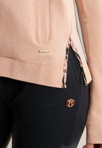 Top rosa claro con una abertura lateral que revela un forro floral, combinado con leggings negros texturizados que presentan un acento de herraje dorado.