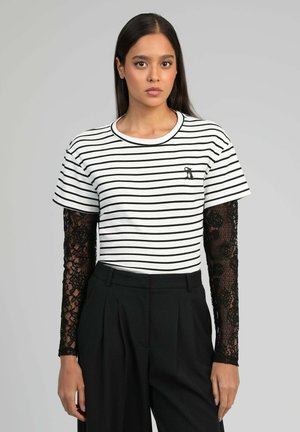 Femme portant une chemise à manches longues en dentelle noire sous une chemise à manches courtes blanche à rayures noires, associée à un pantalon noir taille haute, immobile.