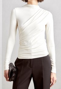 Vit långärmad topp med drapering på sidan, matchad med svarta byxor. Hand som håller en svart vadderad kuvertväska.