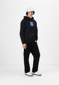 Black hoodie with a blue graphic logo, front pocket, paired with black pants and white sneakers. Light-colored cap completes the outfit.