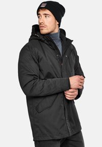 Chaqueta aislante negra con capucha, con cierres de cremallera y botón a presión, bolsillos laterales y un gorro de canalé texturizado. El modelo está de pie con los brazos cruzados.