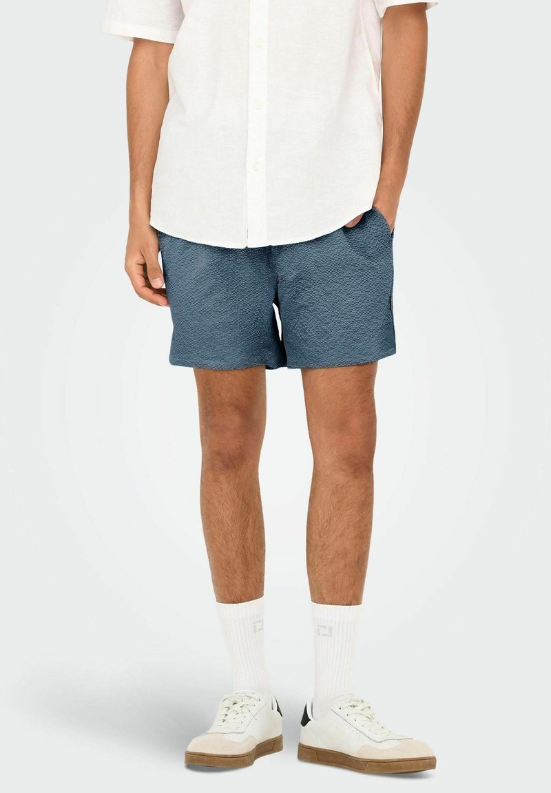 Blue textured shorts with side pockets, paired with a white button-up shirt and white sneakers with brown accents.