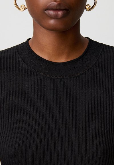 Persona che indossa un top nero a coste con colletto strutturato e grandi orecchini dorati a spirale, mostrata dalle spalle al mento su uno sfondo semplice.