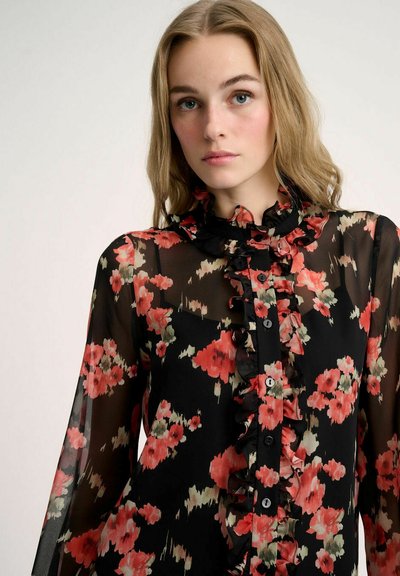 Blusa negra transparente con un patrón floral en rojo y verde, que presenta un cuello con volantes y botón delantero. Mangas largas con puños acampanados.