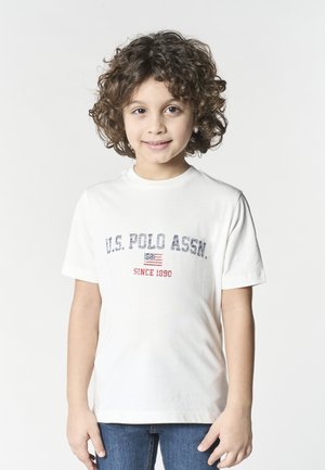 Bambino con capelli ricci che indossa una maglietta bianca U.S. Polo Assn. e jeans blu, in piedi davanti a uno sfondo chiaro e uniforme, sorridendo dolcemente.