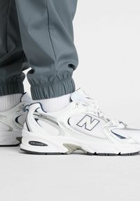 Hvide træningssko har en mesh-overdel med grå detaljer og en polstret pløs. Sålen har et sort skridsikkert mønster. De er sat sammen med grå joggingbukser.