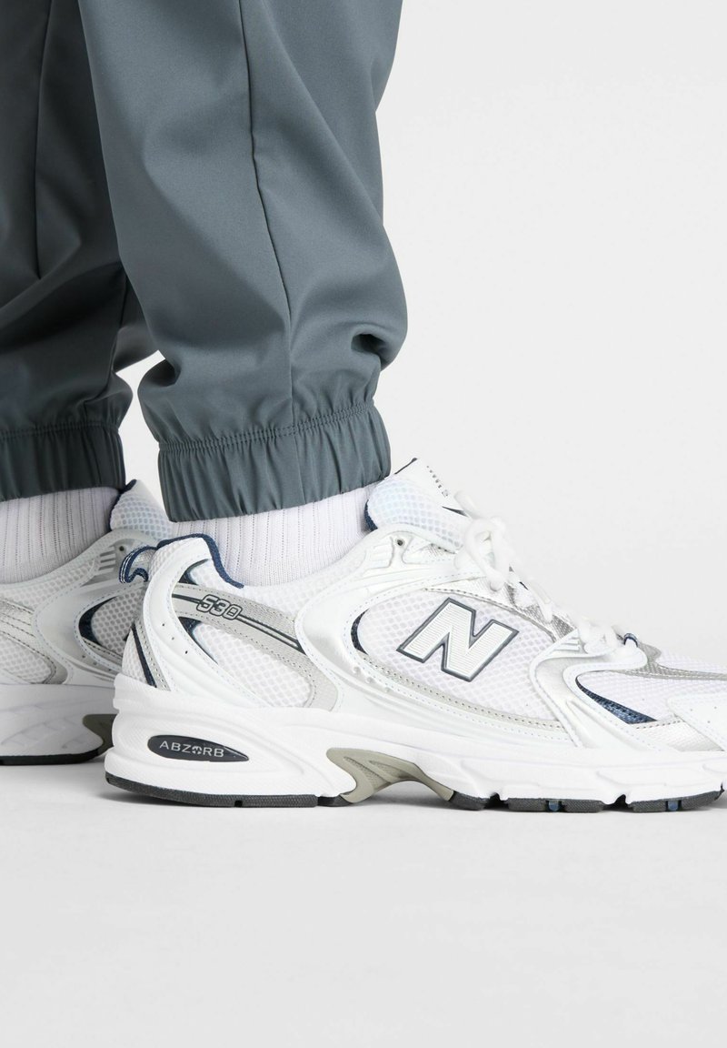 White athletic shoes feature a mesh upper with gray accents and a padded tongue. The sole includes a black traction pattern. Pairing with gray joggers.