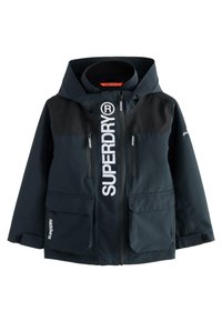 Must ja tumesinine tuulekindel jope lukuga ees, valge ülisuur logo ja neli taskut. Varustatud kapuutsi ja tekstuursete paneelidega.