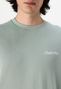 Lys grøn bomulds sweatshirts med rund hals, der har teksten "Club1990" i hvid broderi på fronten. Glat tekstur.