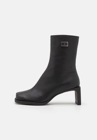 Black ankle boot made of smooth leather, featuring a square toe, a mid-height rectangular heel, and a branded metal accent.
