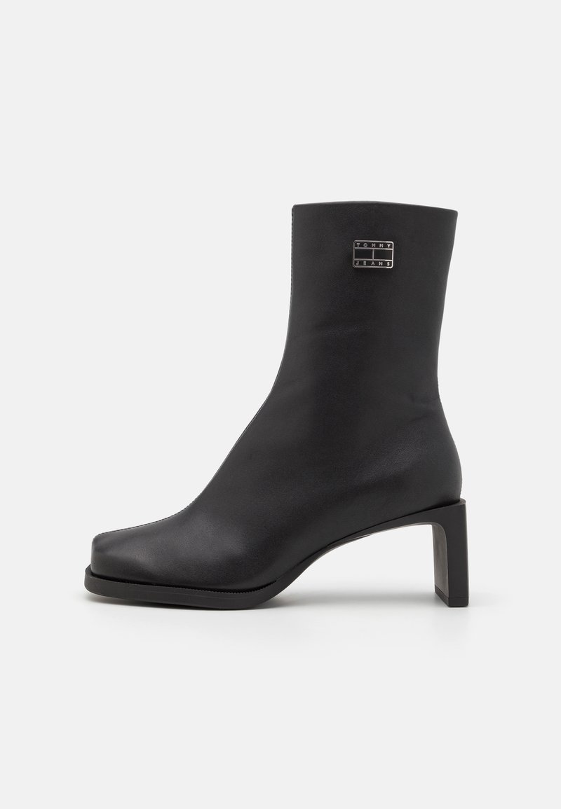 Black ankle boot made of smooth leather, featuring a square toe, a mid-height rectangular heel, and a branded metal accent.