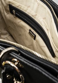 Interiøret af en Guess taske med beige stofforing med gentaget logo, sort lynlåslomme og hardware-detaljer i guldfarve.