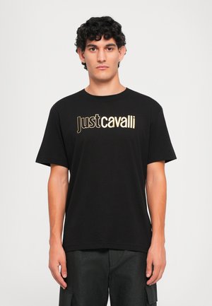 Jauns vīrietis ar cirtainiem matiem, ģērbies melnā Just Cavalli T-kreklā un melnās kabatbiksēs, stāv pret vienkāršu baltu fona.