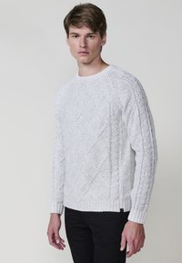 Witte gebreide trui met gestructureerde diamantpatronen en ribgebreide manchetten. Klassiek ontwerp met ronde hals. G gedragen met zwarte broek.
