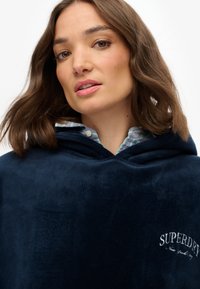 Sudadera de terciopelo azul marino con una gran capucha, que presenta un logo blanco de "SUPERDRY" en el pecho y un acabado texturizado. Debajo hay un cuello a cuadros multicolor.