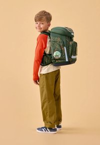 Mochila verde con patrones de hojas, compartimentos con cremallera y acentos reflectantes, llevada por un niño con una camiseta de manga larga roja y crema.