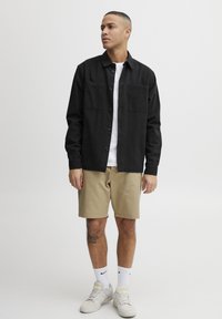 Zwart overhemd met knopen en twee borstzakken, gedragen over een witte t-shirt, gecombineerd met lichtbeige shorts en witte sneakers. Casual ontwerp.